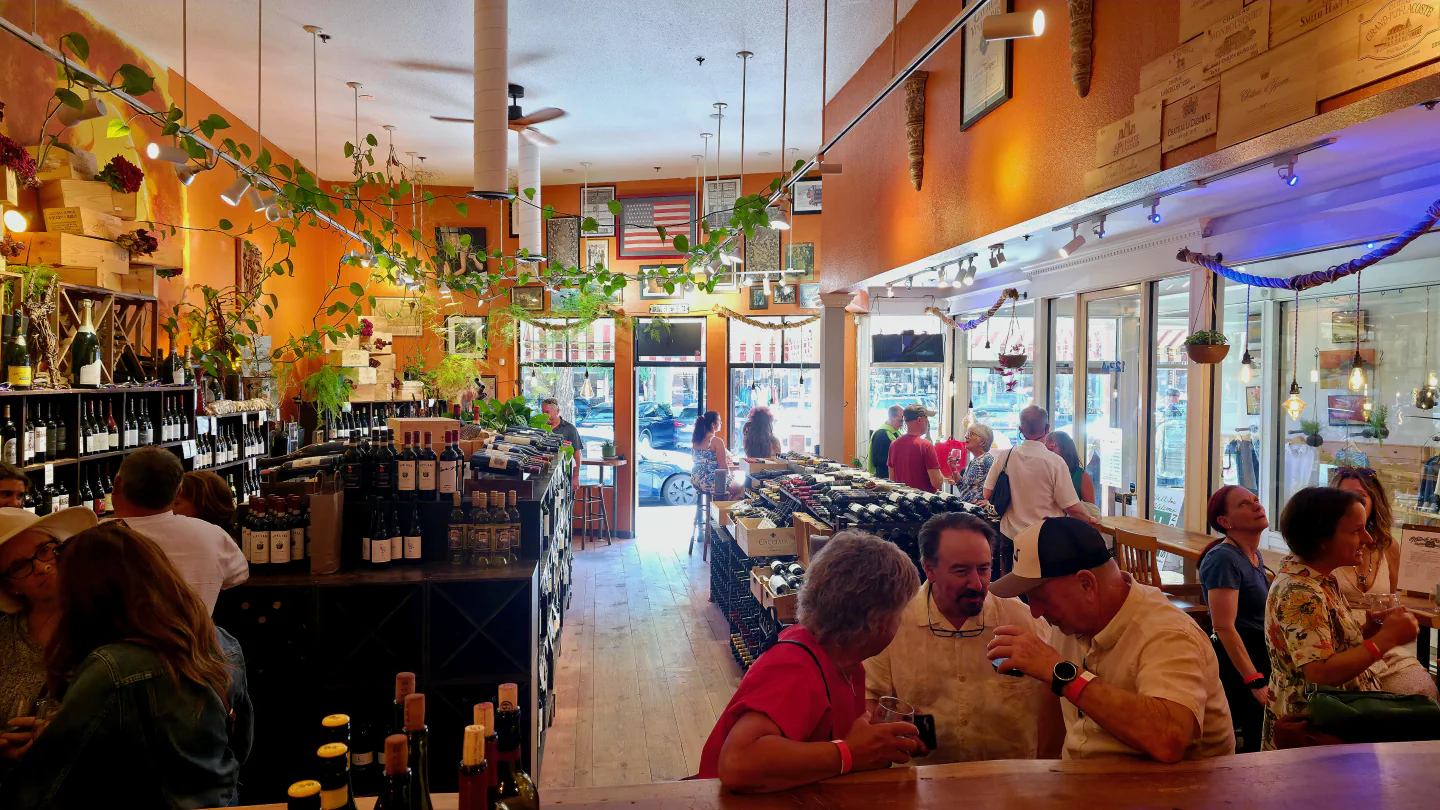 A lively afternoon inside Vine & Barrel, seen from behind the bar with neighbors gathered among the wine shelves